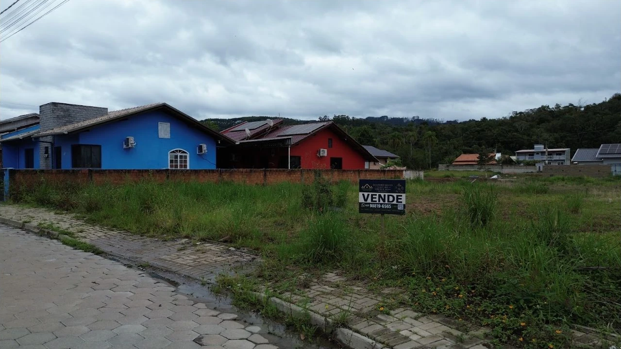 Vista do terreno ao nível da rua, com placa de venda e calçamento em frente