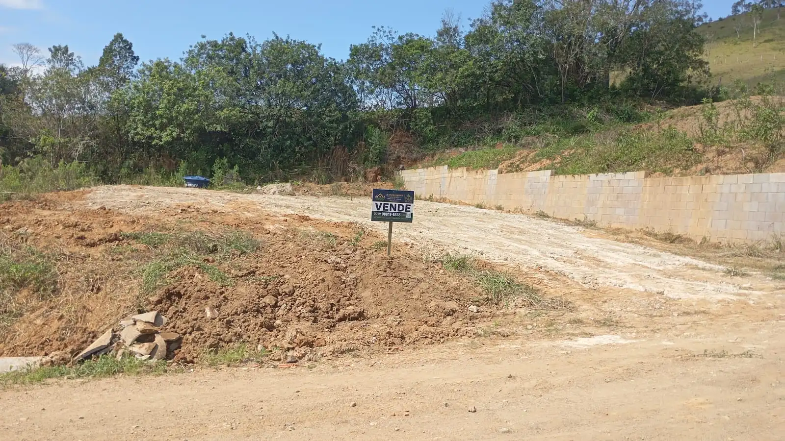 Vista do lote ao nível do terreno com muro lateral e placa de venda